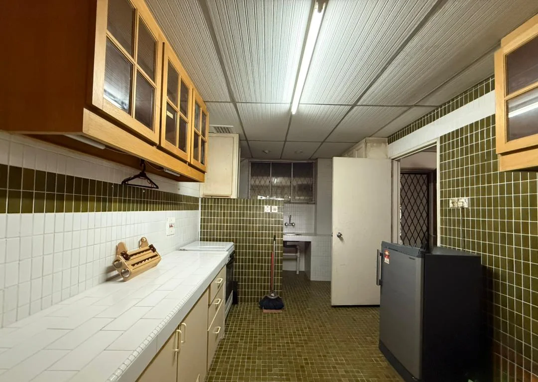 Kitchen features: white tiled countertop, brown wooden cabinetry with glass-front upper cabinets, green tiled backsplash, green tiled flooring, single-bowl stainless-steel sink, chrome faucet, freestanding fridge, door leading to utility area, hanging rack for utensils, overhead fluorescent lighting, ventilation grill, window with security grille.
